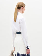 Load image into Gallery viewer, A woman wears the Naroa Blouse—a white top with floral appliqué details—paired with a white Italian-fabric skirt, standing against a plain light background.