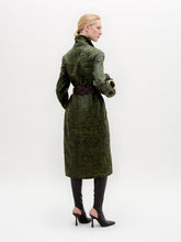 Load image into Gallery viewer, A woman wears the Nilo Trench, a textured dark green coat crafted from Italian fabric, paired with dark boots, a large gold belt buckle, and holding a small dark clutch against a plain background.
