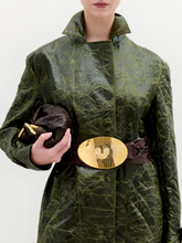 Load image into Gallery viewer, A woman wears the Nilo Trench, a textured dark green coat crafted from Italian fabric, paired with dark boots, a large gold belt buckle, and holding a small dark clutch against a plain background.