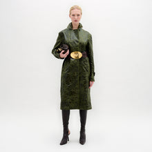 Load image into Gallery viewer, A woman wears the Nilo Trench, a textured dark green coat crafted from Italian fabric, paired with dark boots, a large gold belt buckle, and holding a small dark clutch against a plain background.