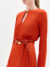 Load image into Gallery viewer, Woman wearing the Pesaro Dress, a long-sleeved orange midi dress with a belted waist crafted from premium Italian fabric, paired with a small purple bag and transparent high-heeled sandals, against a plain white background.
