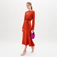 Load image into Gallery viewer, Woman wearing the Pesaro Dress, a long-sleeved orange midi dress with a belted waist crafted from premium Italian fabric, paired with a small purple bag and transparent high-heeled sandals, against a plain white background.
