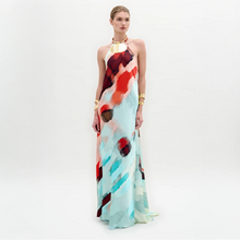 Load image into Gallery viewer, A woman poses against a white background wearing the Zeffa Dress, a sleeveless, floor-length silk maxi with an abstract red, aqua, and white pattern, paired with gold jewelry.