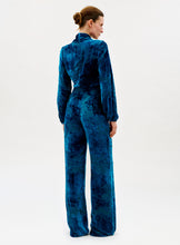Load image into Gallery viewer, A woman faces away, wearing the high-waisted Como Pant in blue velvet with wide legs. Her hair is tied back in a neat bun as she poses against a plain, light background.
