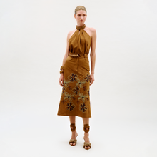 Load image into Gallery viewer, A woman stands against a plain background wearing the Layan Skirt, styled with brown sandals featuring floral accents, a matching belt, and bracelets. Her hair is pulled back in a sleek style.
