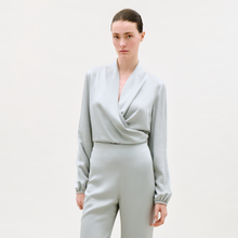 Load image into Gallery viewer, A woman wearing the Tea Blouse in light gray with long sleeves and wrap style, paired with matching high-waisted pants, stands against a plain light background, gazing confidently at the camera.
