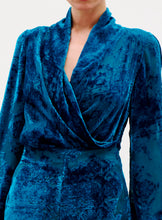 Load image into Gallery viewer, A person wearing the Tea Blouse, made from luxurious Italian blue crushed velvet with long sleeves and a deep V-neckline. The blouse has a soft, textured look with intricate patterns, set against a plain, light-colored background.
