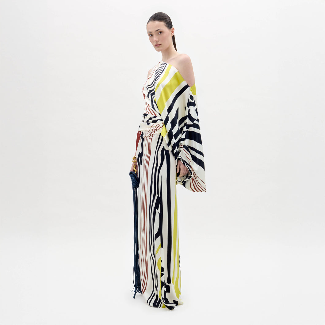 A woman stands in a white space wearing the Dotty Dress—a one-shoulder, floor-length gown with bold black, yellow, and red stripes—while holding a tasseled accessory.