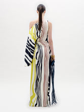 Load image into Gallery viewer, A woman stands in a white space wearing the Dotty Dress—a one-shoulder, floor-length gown with bold black, yellow, and red stripes—while holding a tasseled accessory.
