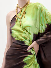 Load image into Gallery viewer, A person models the Afrodita Dress—a one-shoulder, floor-length gown with a green and brown tie-dye print—paired with gold necklaces, against a plain white background.
