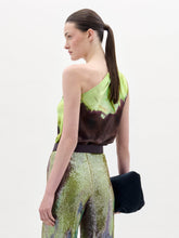 Load image into Gallery viewer, A woman stands against a plain background wearing the Yanet Blouse, a one-shoulder green and brown top, paired with sequined pants, a wide belt with a large buckle, and holding a black clutch.
