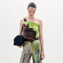 Load image into Gallery viewer, A woman stands against a plain background wearing the Yanet Blouse, a one-shoulder green and brown top, paired with sequined pants, a wide belt with a large buckle, and holding a black clutch.
