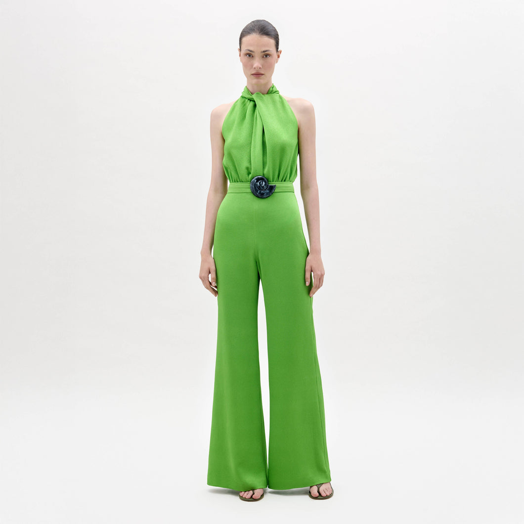 A woman stands facing forward in the Siv Jumpsuit, a bright green halter-neck style with wide-leg pants and a matching belt, paired with black sandals, against a plain white background.