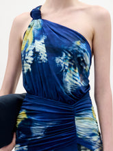 Load image into Gallery viewer, A woman stands against a plain white background wearing the Antara Dress, a one-shoulder, floor-length blue gown with yellow and white patterns, holding a black clutch.
