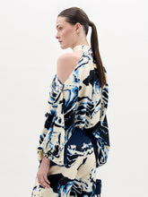 Load image into Gallery viewer, A woman stands against a plain background wearing the Safira Blouse, a high-neck, cold-shoulder top with matching wide-leg pants in a blue, black, and white marble pattern.
