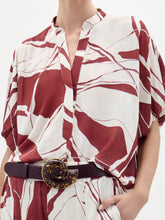Load image into Gallery viewer, A woman poses against a plain white background wearing the Susanne Blouse, featuring a red and white abstract pattern, matched with coordinating pants and accessorized with a black belt with a gold buckle.

