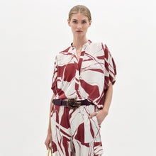 Load image into Gallery viewer, A woman poses against a plain white background wearing the Susanne Blouse, featuring a red and white abstract pattern, matched with coordinating pants and accessorized with a black belt with a gold buckle.
