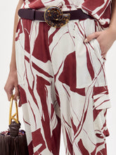 Load image into Gallery viewer, A woman stands against a plain background wearing the Jess Pant in a red and white pattern, gold sandals, a dark belt, and holding a dark fringed bag.
