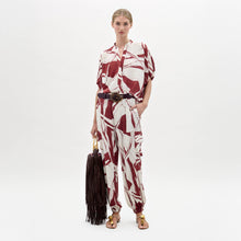 Load image into Gallery viewer, A woman stands against a plain background wearing the Jess Pant in a red and white pattern, gold sandals, a dark belt, and holding a dark fringed bag.
