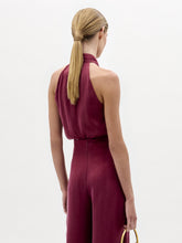 Load image into Gallery viewer, A woman stands against a plain background wearing the Shela Blouse, a sleeveless, high-neck magenta top with a tie detail at the waist, paired with a small handbag.
