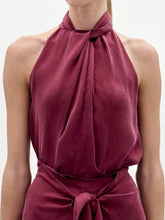Load image into Gallery viewer, A woman stands against a plain background wearing the Shela Blouse, a sleeveless, high-neck magenta top with a tie detail at the waist, paired with a small handbag.
