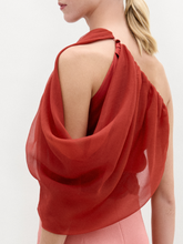 Load image into Gallery viewer, A woman wears the Adil Blouse in red with a one-shoulder neckline, paired with pink high-waisted pants, a wide belt with a large gold buckle, and gold bracelets, standing against a plain white background.
