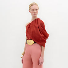 Load image into Gallery viewer, A woman wears the Adil Blouse in red with a one-shoulder neckline, paired with pink high-waisted pants, a wide belt with a large gold buckle, and gold bracelets, standing against a plain white background.
