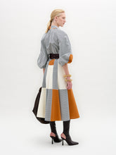 Load image into Gallery viewer, A woman stands against a white background wearing the Agostina Dress—striped and color-blocked, crafted from Italian fabric—paired with high brown boots, a wide belt, and holding a large dark bag.
