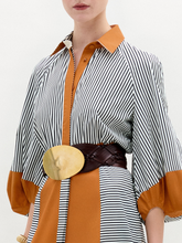 Load image into Gallery viewer, A woman stands against a white background wearing the Agostina Dress—striped and color-blocked, crafted from Italian fabric—paired with high brown boots, a wide belt, and holding a large dark bag.
