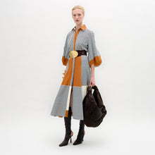 Load image into Gallery viewer, A woman stands against a white background wearing the Agostina Dress—striped and color-blocked, crafted from Italian fabric—paired with high brown boots, a wide belt, and holding a large dark bag.
