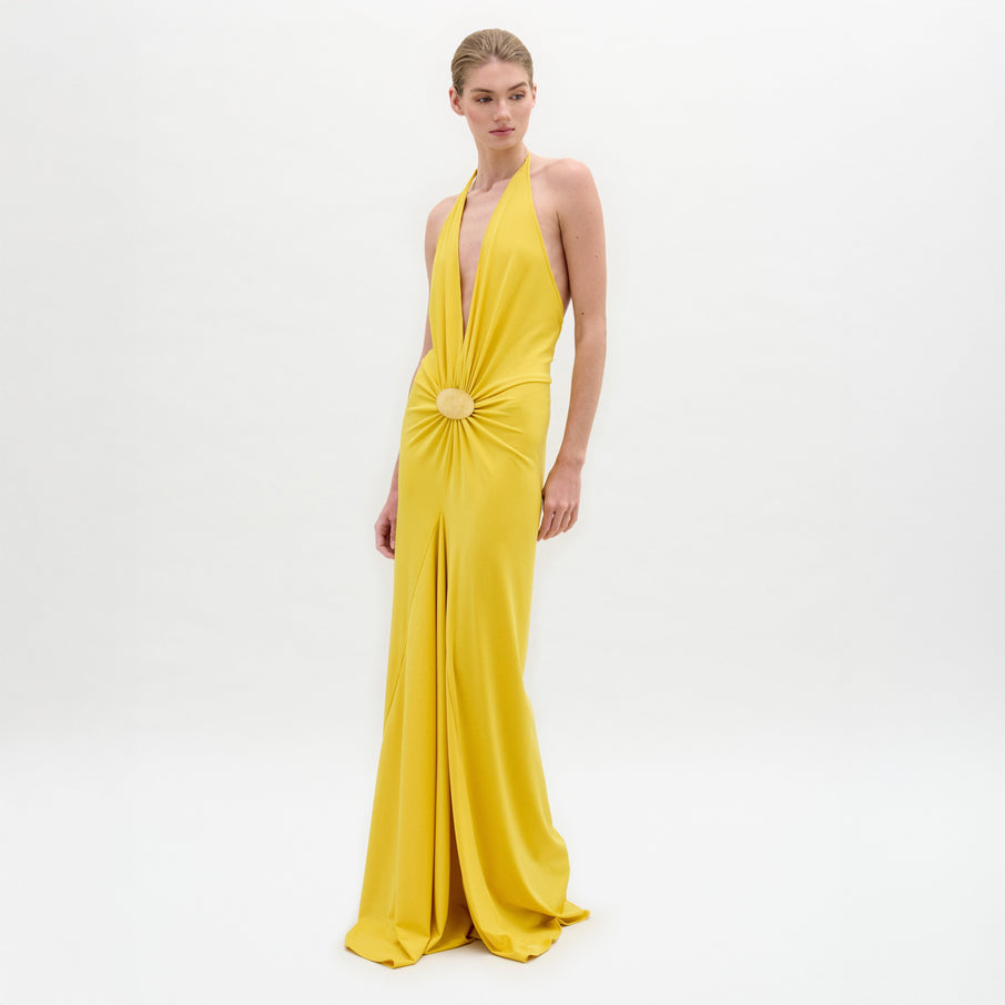 Model in the Aiko Dress—a floor-length, sleeveless yellow maxi with a deep halter neckline and gathered circular waist detail—stands against a plain white background.
