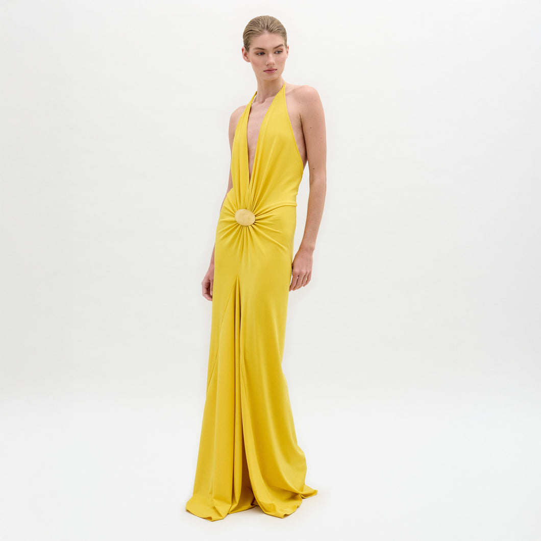 Model in the Aiko Dress—a floor-length, sleeveless yellow maxi with a deep halter neckline and gathered circular waist detail—stands against a plain white background.