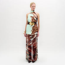 Load image into Gallery viewer, A woman poses against a plain backdrop in the Amalia Skirt—a flowing maxi with an abstract brown and blue pattern—paired with a sleeveless halter top, wide black belt, and bracelets. The look features luxurious Italian fabric.
