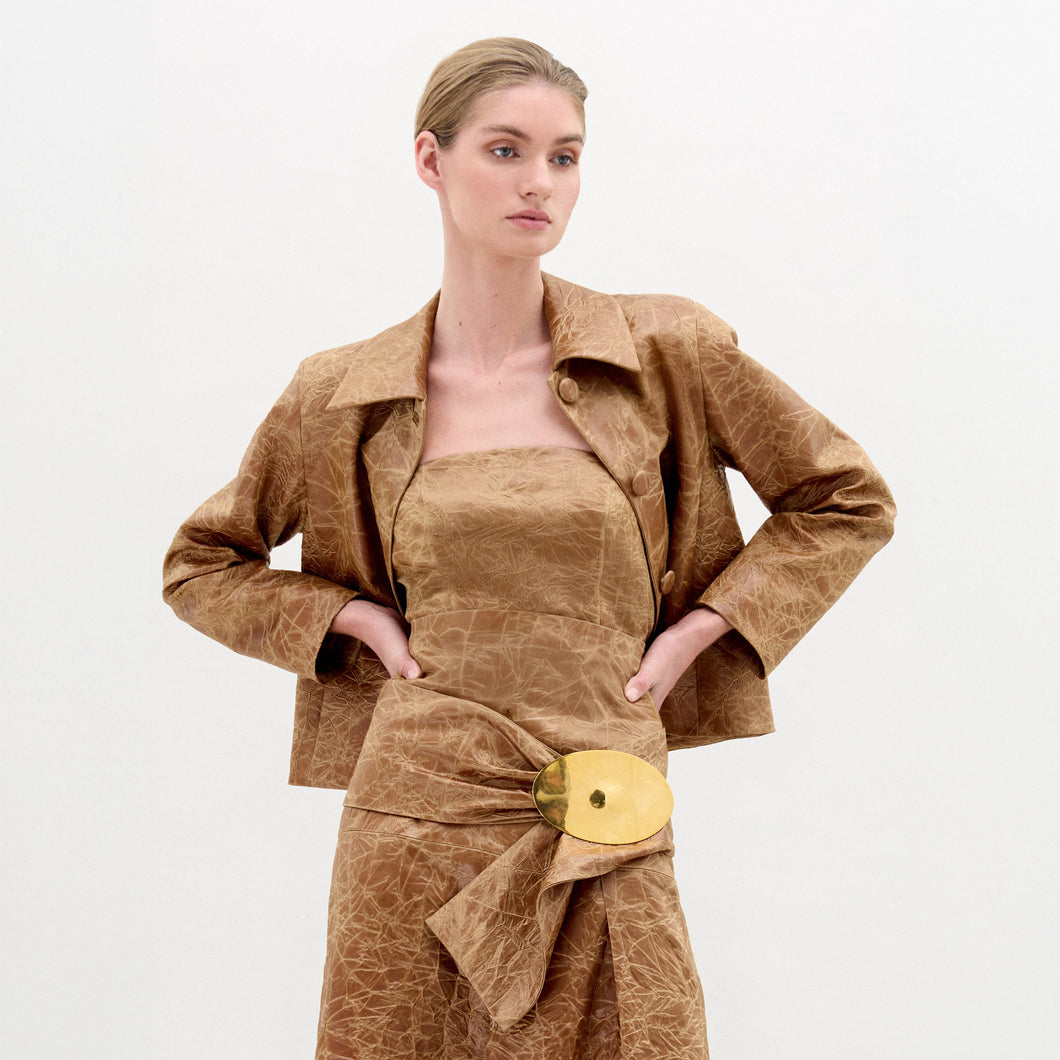 A woman wearing the Astor Top, a crinkled brown strapless style with a fitted waist and large gold belt buckle, stands against a plain white background.