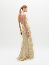 Load image into Gallery viewer, A woman wears the Aurora Dress—floor-length, sleeveless, crafted from gold lace with a fitted silhouette and wavy patterns—paired with gold cuff bracelets, standing against a plain white background.

