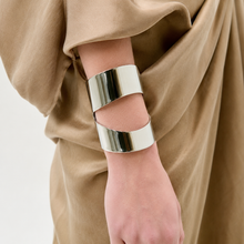 Load image into Gallery viewer, A person wears a camel-colored draped garment and the Artemis Cuff—a wide, modern silver bracelet with a unique overlapping open design—on their wrist.
