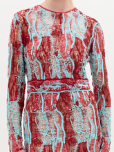 Load image into Gallery viewer, A person stands against a plain background wearing the Barisa Dress, a long, patterned red and blue fitted dress, and holding a small purple handbag.
