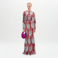 Load image into Gallery viewer, A person stands against a plain background wearing the Barisa Dress, a long, patterned red and blue fitted dress, and holding a small purple handbag.
