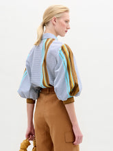 Load image into Gallery viewer, A person stands against a plain background wearing the oversized fit Belkis Blouse with balloon sleeves and stripes, paired with tan high-waisted pants, and holding a tan accessory in their left hand.
