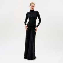 Load image into Gallery viewer, A woman wears the Birdy Dress—a long-sleeved, floor-length black gown featuring a small front keyhole cutout and gathered waist—standing against a plain, light background.
