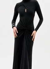 Load image into Gallery viewer, A person wearing the Birdy Dress, a long-sleeved black dress featuring a keyhole chest cutout and gathered twist at the waist, with a bracelet on one wrist, stands against a plain light background.
