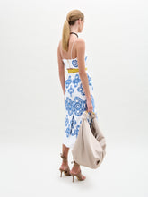 Load image into Gallery viewer, A woman stands against a plain background wearing the Ceres Dress, an embroidered linen midi with blue patterns and a yellow belt, crafted from fine Italian fabric. She pairs it with tan sandals and carries a beige shoulder bag.

