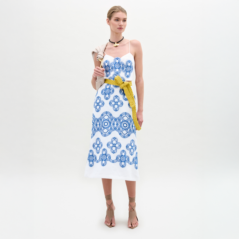 A woman stands against a plain background wearing the Ceres Dress, an embroidered linen midi with blue patterns and a yellow belt, crafted from fine Italian fabric. She pairs it with tan sandals and carries a beige shoulder bag.