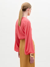 Load image into Gallery viewer, A woman stands against a plain background wearing the Covilha Blouse, a silk coral long-sleeve top, paired with a tan skirt featuring a large gold circular belt detail.
