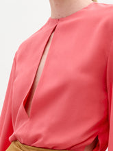 Load image into Gallery viewer, A woman stands against a plain background wearing the Covilha Blouse, a silk coral long-sleeve top, paired with a tan skirt featuring a large gold circular belt detail.
