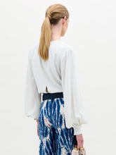 Load image into Gallery viewer, A woman poses against a plain background wearing the Covilha Blouse, featuring a keyhole neckline and balloon sleeves, styled with high-waisted blue patterned pants and holding a small beige bag.
