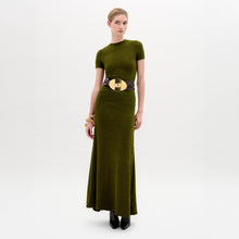 Load image into Gallery viewer, A woman wearing the Cristal Dress, an olive green slim fit maxi dress paired with a wide black belt with a gold buckle, black boots, and gold bracelets stands against a plain white background.
