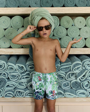 Load image into Gallery viewer, Bo wearing sunglasses and swim shorts standing in front of shelves with folded towels.
