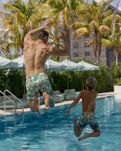 Load image into Gallery viewer, Man and boy in floral swim trunks jumping into a pool with palm trees and a building in the background.
