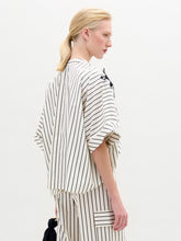 Load image into Gallery viewer, A person with blond hair in a low ponytail wears the Daia Blouse, a loose, white 100% cotton top with black vertical stripes and short sleeves, standing against a plain light background.
